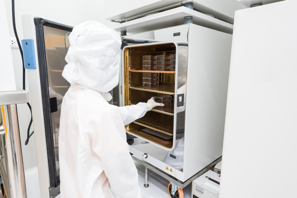 A scientist in sterile coverall gown placing cell culture flasks in the CO2 incubator. Doing biological research in clean environment. Cleanroom facility